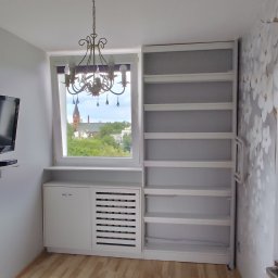 Michał Przybyszewski - Biała szafa wnękowa z otwartymi półkami i zabudową grzejnika, widok na miasto z kościołem w tle przez okno z ozdobnym żyrandolem.