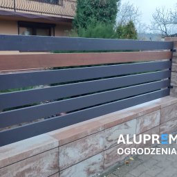 ALUPREM ZBIGNIEW OLCZAK - Nowoczesne ogrodzenie panelowe z szarymi, poziomymi przęsłami i elementami imitującymi drewno, osadzone na murku z bloczków w odcieniach beżu i brązu, na pierwszym planie widoczny napis...