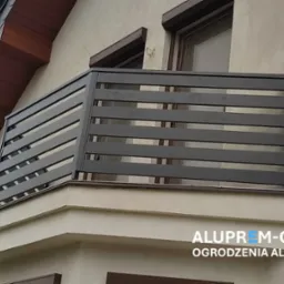 Nowoczesna balustrada balkonowa z poziomych, grafitowych profili aluminiowych, zamontowana na balkonie budynku z beżową elewacją i brązowymi elementami wykończeniowymi