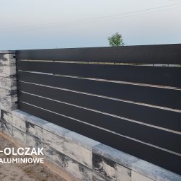 ALUPREM ZBIGNIEW OLCZAK - Nowoczesne, poziome przęsła w kolorze antracytu zamontowane na słupkach z szarej, marmurkowej kostki, widok na linię horyzontu i pojedyncze drzewo w oddali.