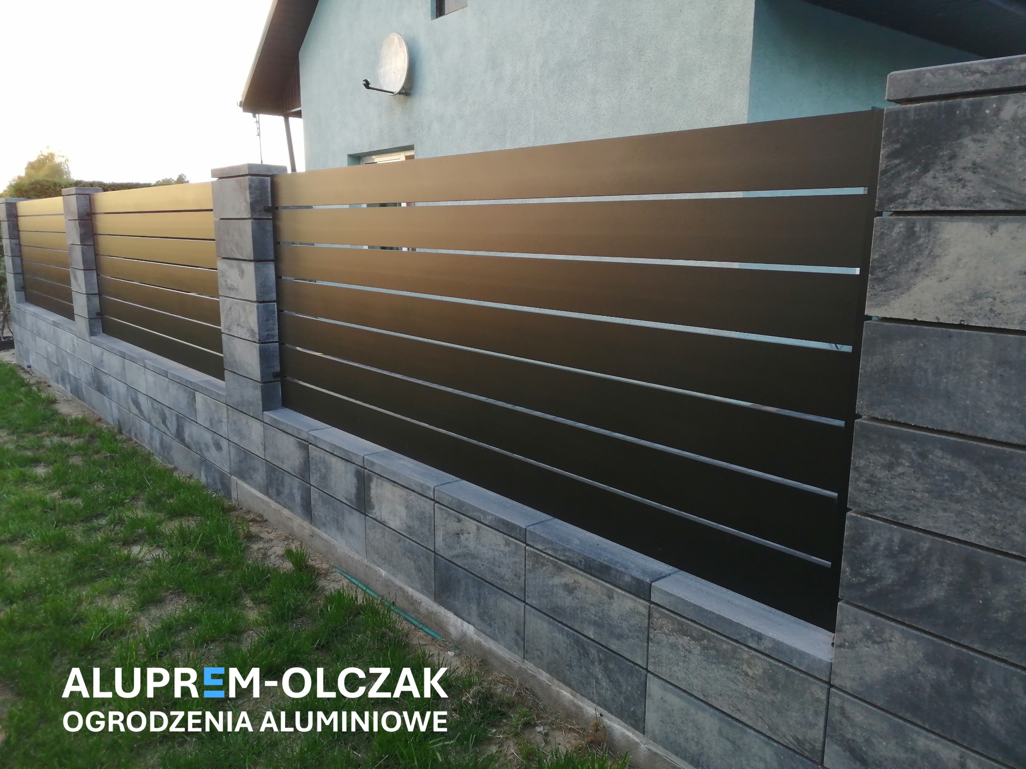 Nowoczesne, poziome przęsła ogrodzeniowe w kolorze antracytowym, osadzone na szarych, betonowych słupkach i podmurówce, na tle trawnika i jasnego domu.