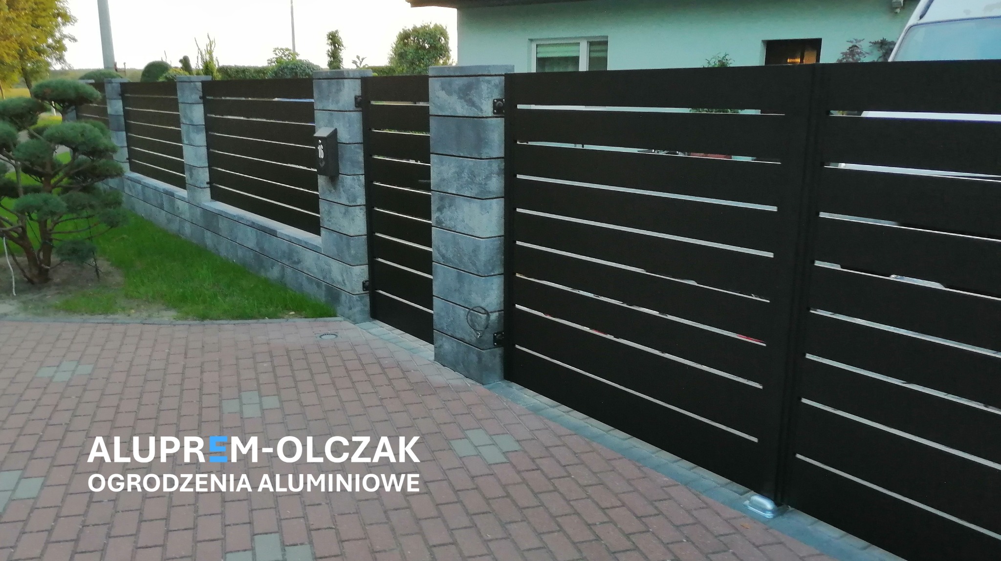 Nowoczesne, poziome, czarne przęsła ogrodzeniowe z jasnymi prześwitami, osadzone na szarych, kwadratowych słupkach z betonu architektonicznego, widok z zewnątrz posesji na tle zieleni i domu.