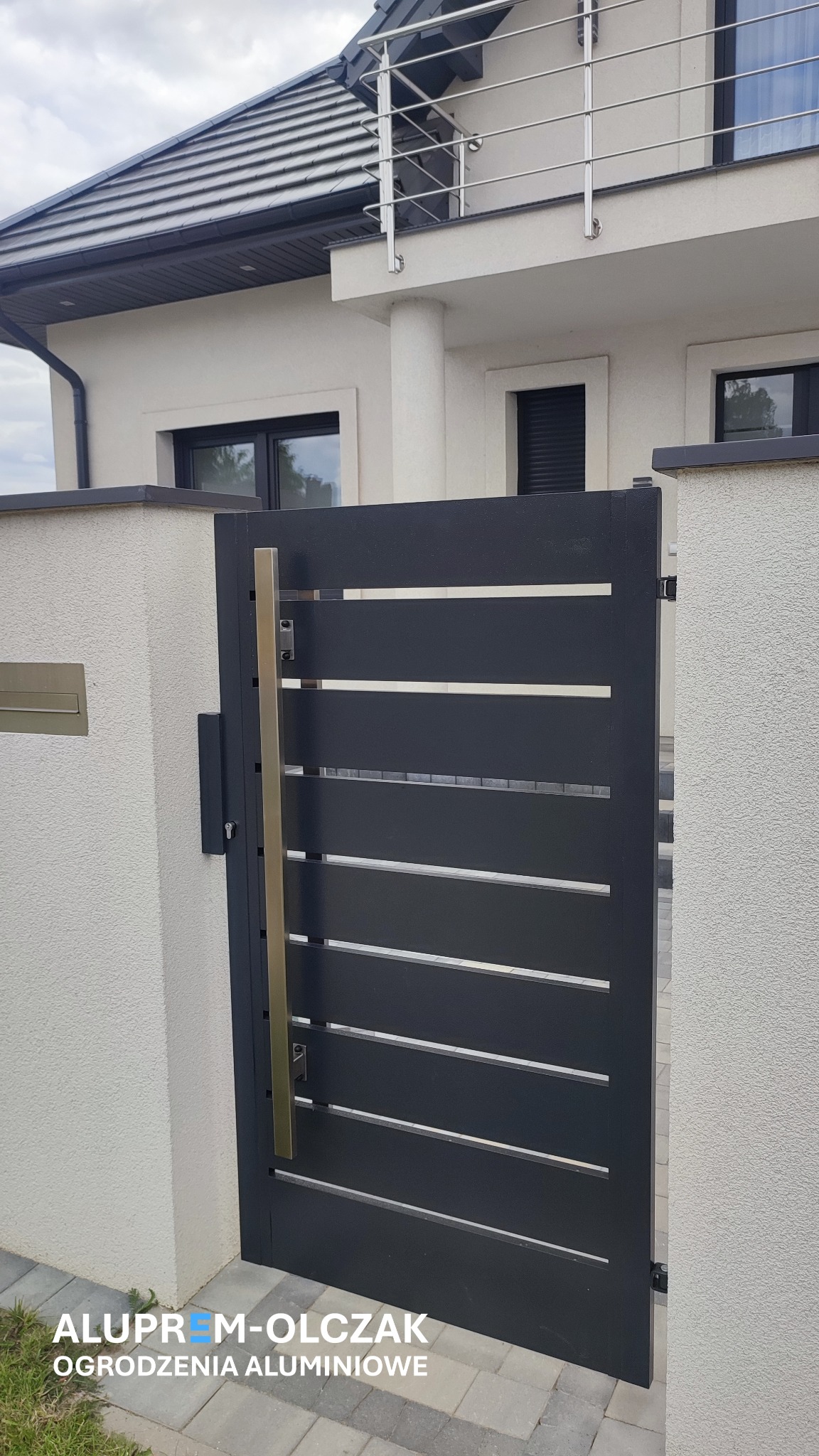 Nowoczesna, antracytowa brama z poziomymi szczeblami i złotym uchwytem, zamontowana między dwoma słupkami z jasnego tynku strukturalnego, na tle domu z balkonem z balustradą ze stali nierdzewnej.