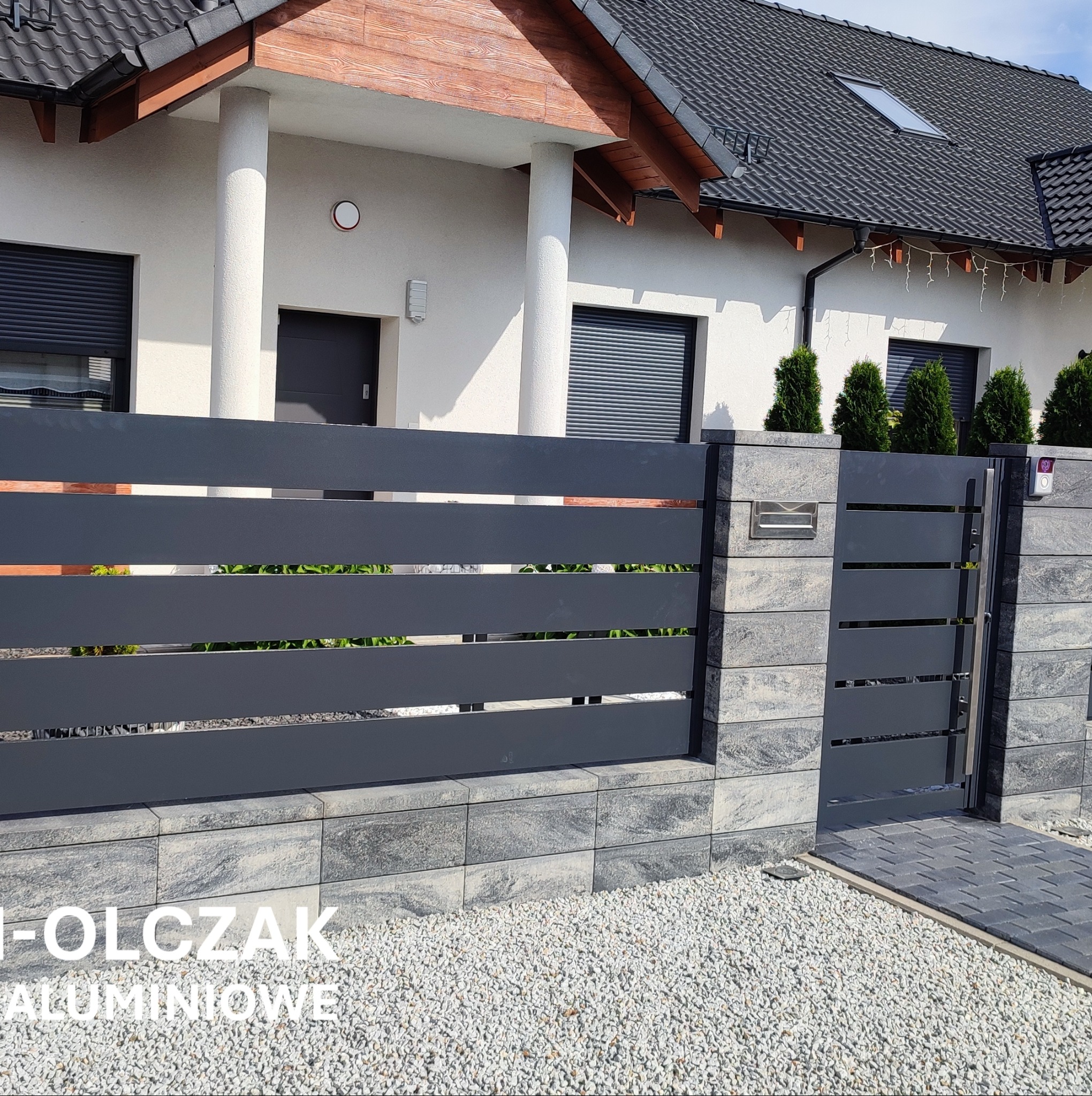 Nowoczesne, poziome przęsła w ciemnoszarej kolorystyce, tworzące ogrodzenie posesji z furtką, uzupełnione kamienną podmurówką i słupkami, na tle jasnego domu z ciemną dachówką i zielenią.
