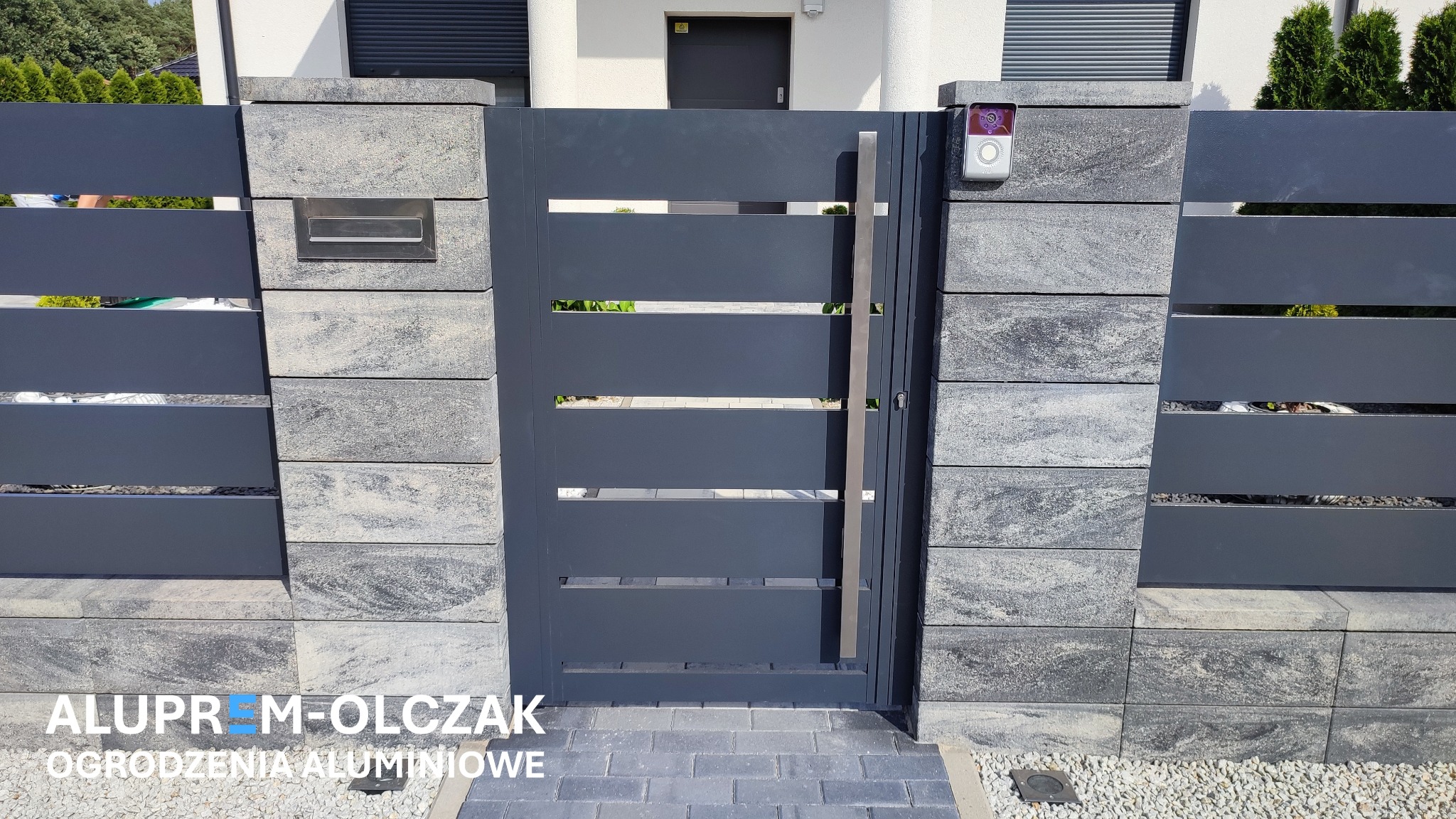 Nowoczesna brama metalowa w kolorze grafitowym, z poziomymi szczelinami i długim, pionowym uchwytem ze stali nierdzewnej, osadzona między słupkami z szarej kostki imitującej kamień, z zamontowanym...