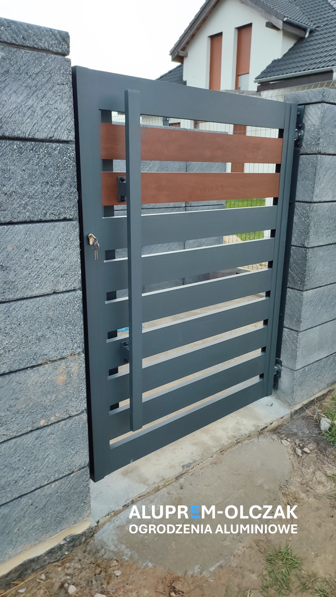 Nowoczesna, grafitowa furtka z poziomymi, drewnopodobnymi wstawkami, zamontowana na słupkach z szarego kamienia łupanego; klucze w zamku.