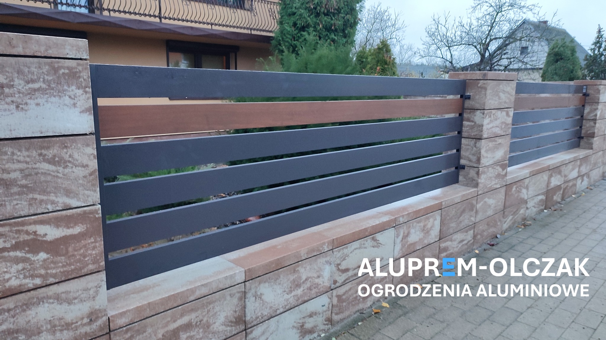 Nowoczesne ogrodzenie panelowe z szarymi, poziomymi przęsłami i elementami imitującymi drewno, osadzone na murku z bloczków w odcieniach beżu i brązu, na pierwszym planie widoczny napis...
