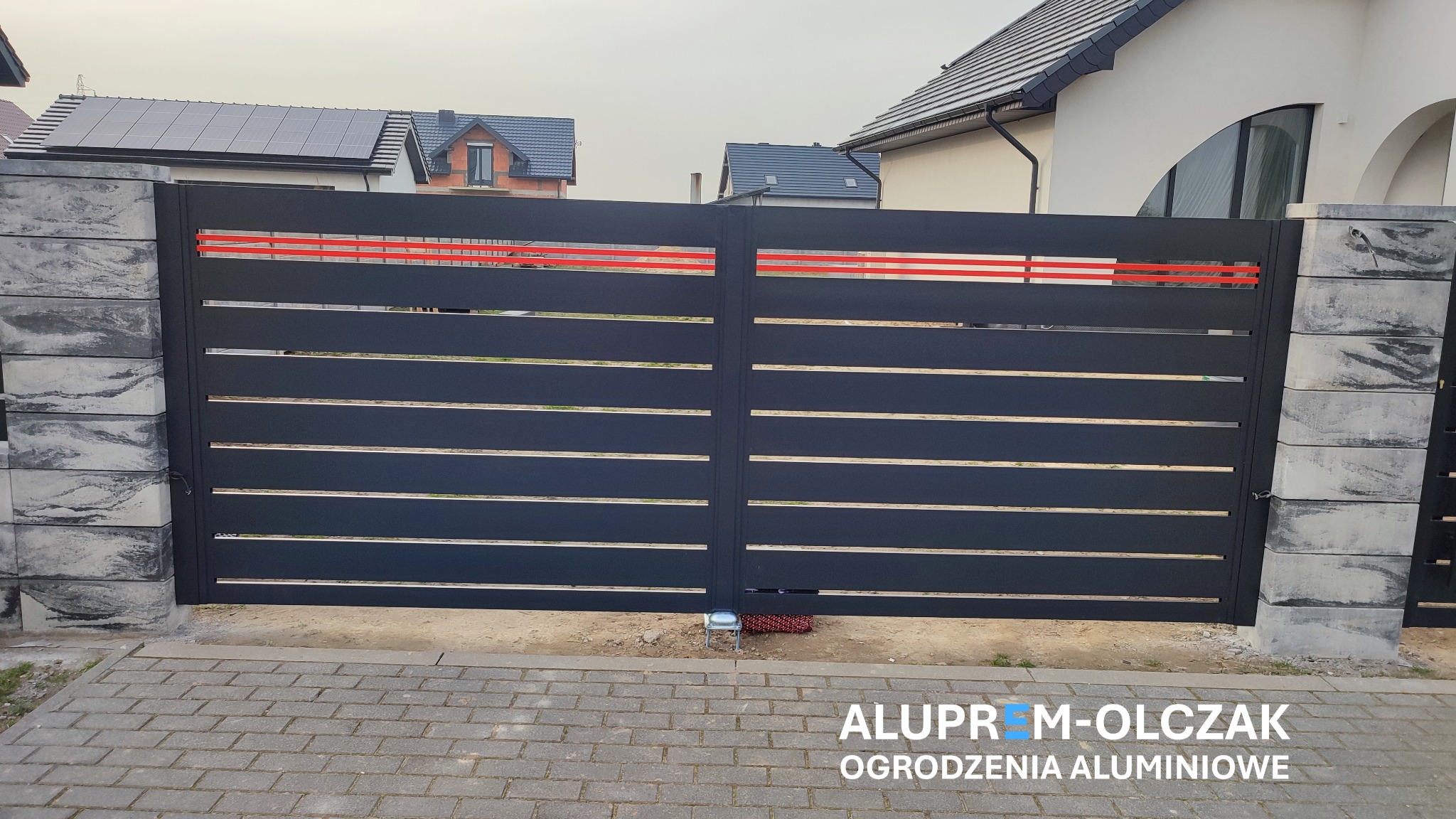 Nowoczesna, przesuwna brama w kolorze antracytowym z poziomymi, srebrnymi profilami, zamontowana pomiędzy dwoma słupkami z szarego, łupanego betonu, z widocznymi domami w tle.