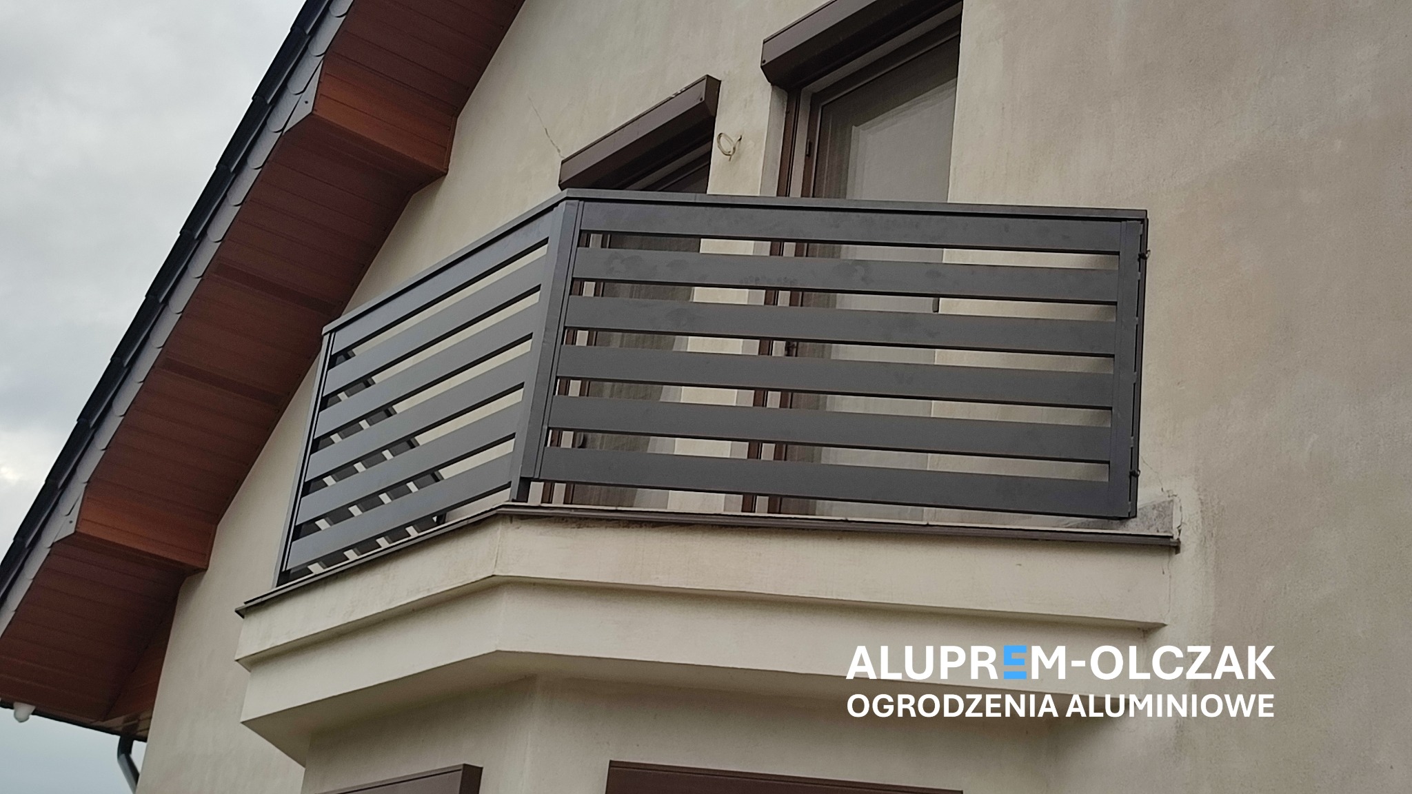 Nowoczesna balustrada balkonowa z poziomych, grafitowych profili aluminiowych, zamontowana na balkonie budynku z beżową elewacją i brązowymi elementami wykończeniowymi