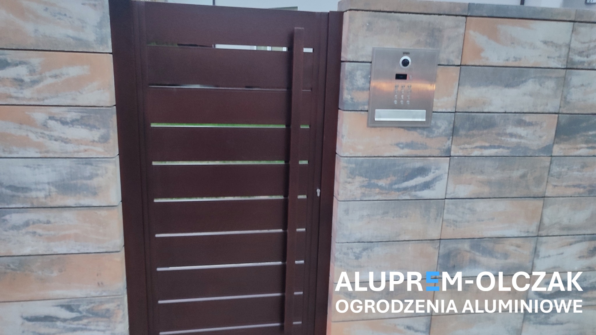 Nowoczesna brama z poziomych profili w kolorze ciemnego brązu, zamontowana na słupku z dekoracyjnych betonowych bloczków, z widocznym domofonem.