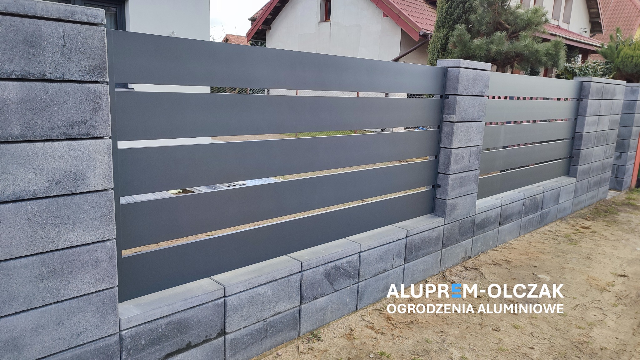 Nowoczesne ogrodzenie z poziomych, szarych paneli aluminiowych na podmurówce z szarych bloczków betonowych, z widocznym logo firmy na dole zdjęcia.