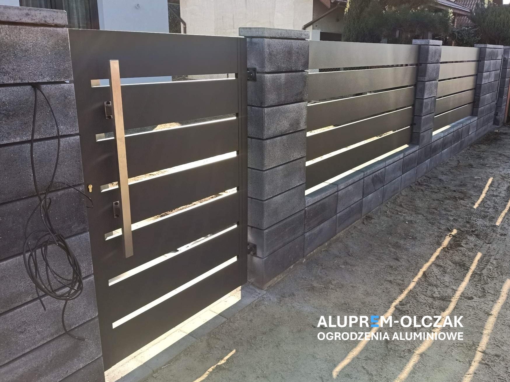 Nowoczesna, pozioma brama wjazdowa z szarego metalu z długim, prostokątnym uchwytem, osadzona w słupkach z szarej kostki betonowej, z widocznymi kablami przy zamku.