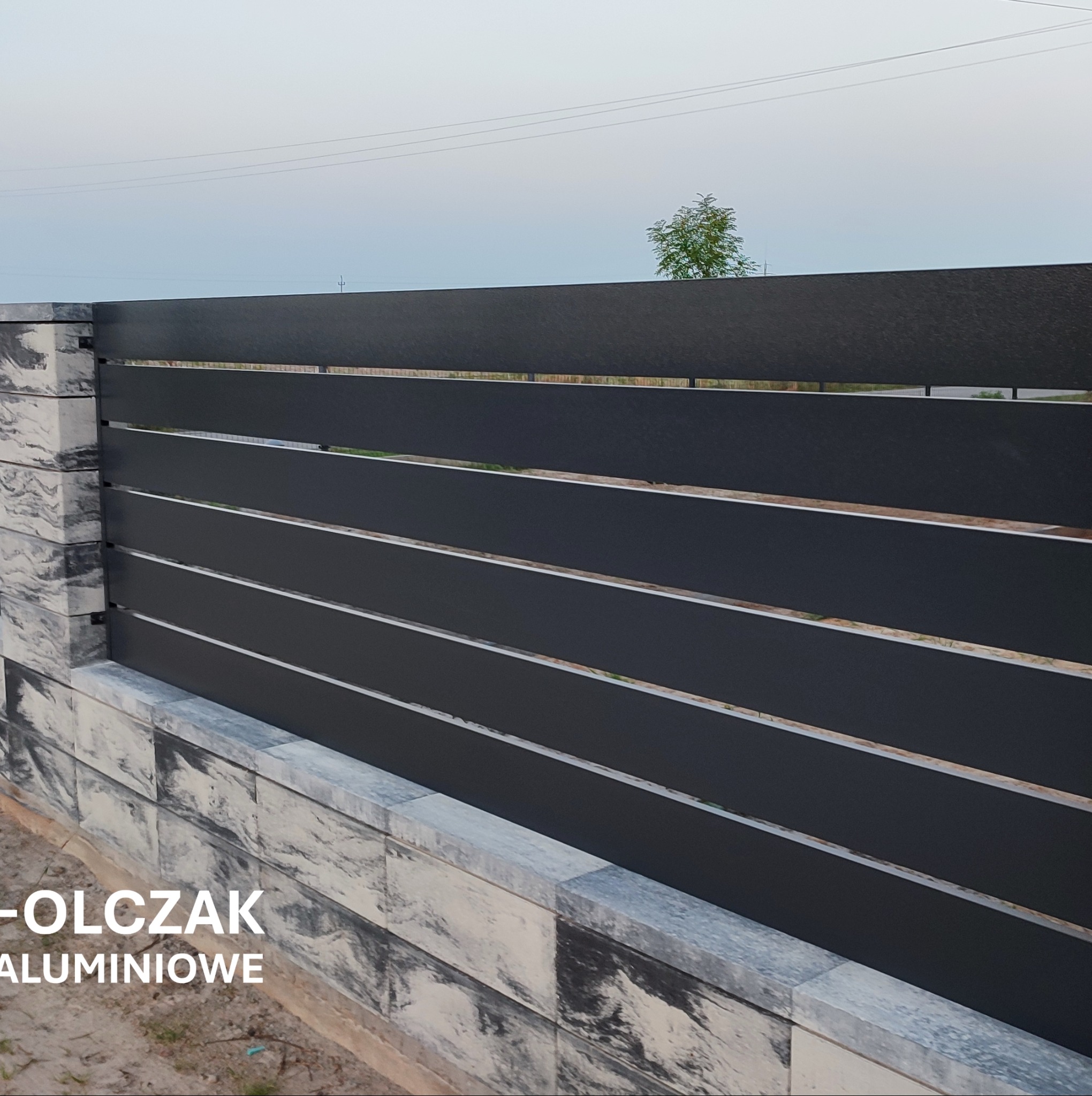 Nowoczesne, poziome przęsła w kolorze antracytu zamontowane na słupkach z szarej, marmurkowej kostki, widok na linię horyzontu i pojedyncze drzewo w oddali.