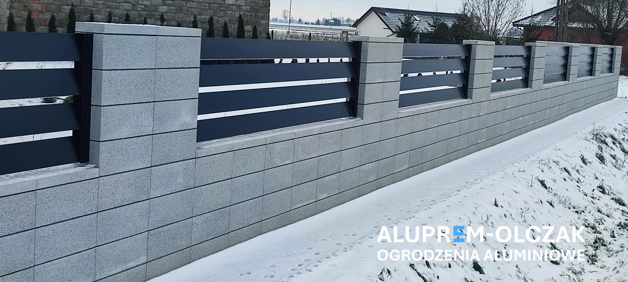 Nowoczesne ogrodzenie z szarej, betonowej podmurówki i czarnych, poziomych paneli, widok z zewnątrz w zimowej scenerii.