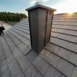 Kamil Sarosiak - Nowoczesny, ciemnoszary komin wentylacyjny z metalowym daszkiem na dachu z ciemnej dachówki ceramicznej, widok z góry, w tle zieleń drzew i błękitne niebo z chmurami, promienie zachodzącego słońca.