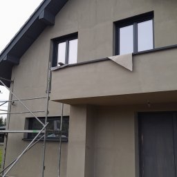 Kamil Sarosiak - Dom jednorodzinny w trakcie prac elewacyjnych, widoczne rusztowanie, ocieplenie fundamentów i zabezpieczony balkon z fragmentem materiału ochronnego, szara dachówka.