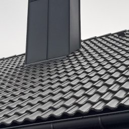 Kamil Sarosiak - Szary komin wentylacyjny z blachy na dachu pokrytym dachówką ceramiczną w odcieniach szarości, widok z dołu na tle pochmurnego nieba.