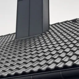 Szary komin wentylacyjny z blachy na dachu pokrytym dachówką ceramiczną w odcieniach szarości, widok z dołu na tle pochmurnego nieba.