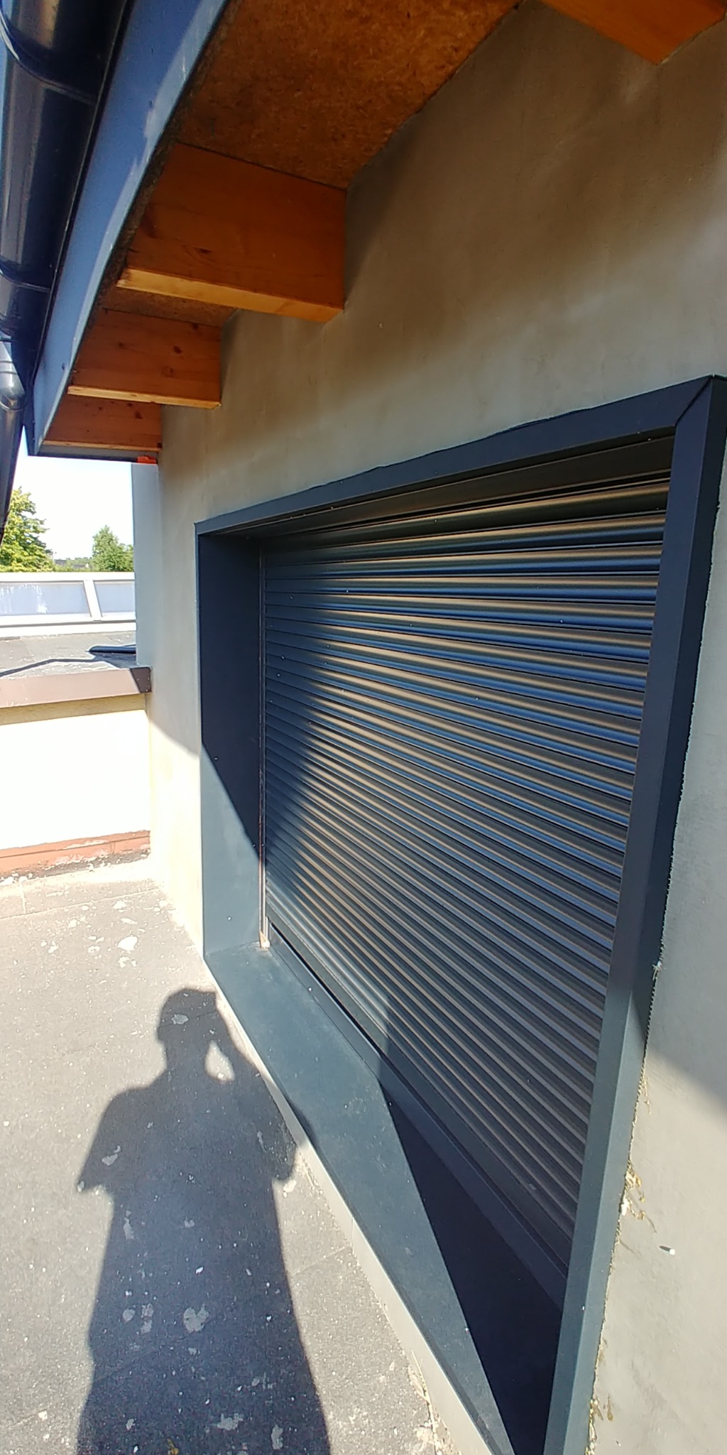 Zamknięta, szara roleta zewnętrzna w oknie pod okapem dachu, widoczny cień osoby robiącej zdjęcie.