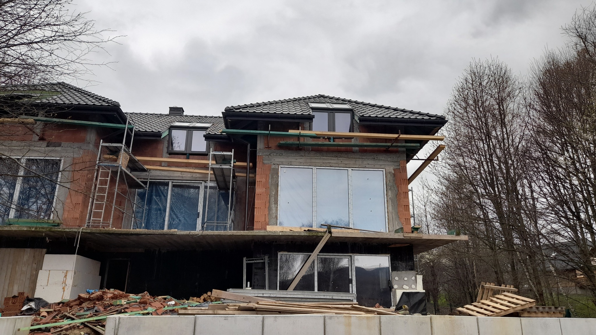 Dom w trakcie budowy, z widocznym fragmentem dachu pokrytego ciemną dachówką, rusztowaniem, niezabudowanymi oknami i elementami konstrukcyjnymi, na tle pochmurnego nieba i drzew bez liści.