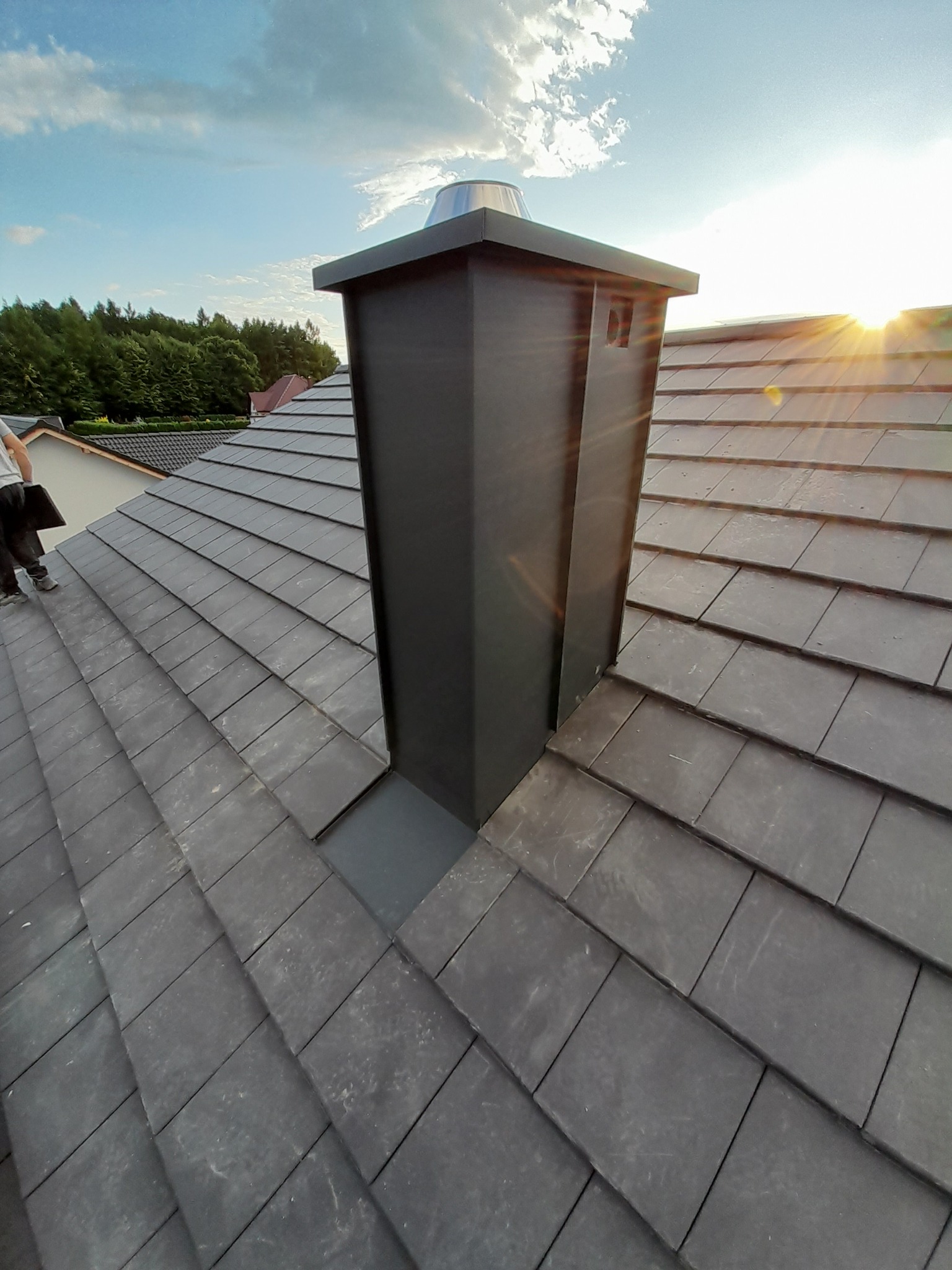 Nowoczesny, ciemnoszary komin wentylacyjny z metalowym daszkiem na dachu z ciemnej dachówki ceramicznej, widok z góry, w tle zieleń drzew i błękitne niebo z chmurami, promienie zachodzącego słońca.