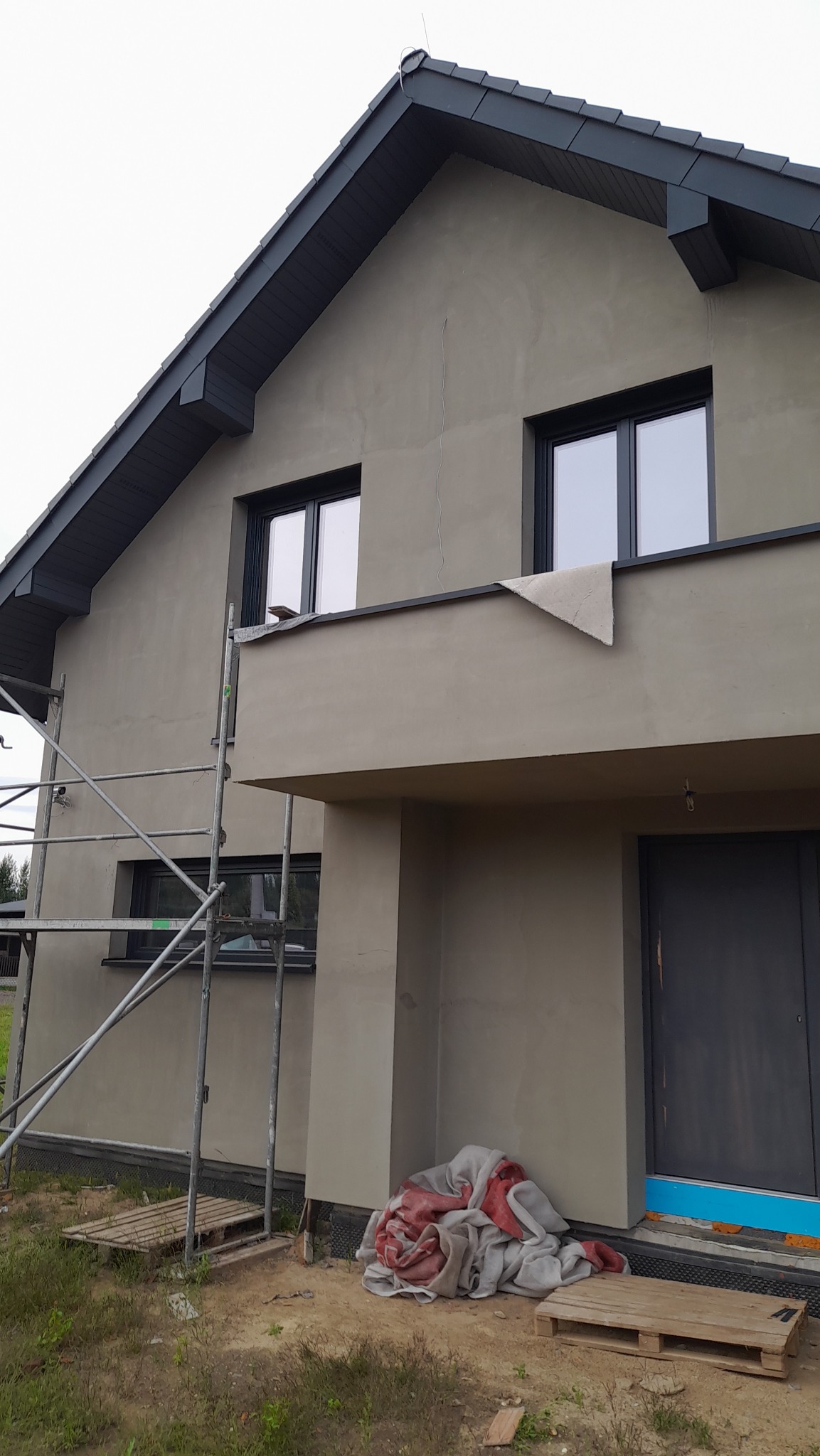 Dom jednorodzinny w trakcie prac elewacyjnych, widoczne rusztowanie, ocieplenie fundamentów i zabezpieczony balkon z fragmentem materiału ochronnego, szara dachówka.