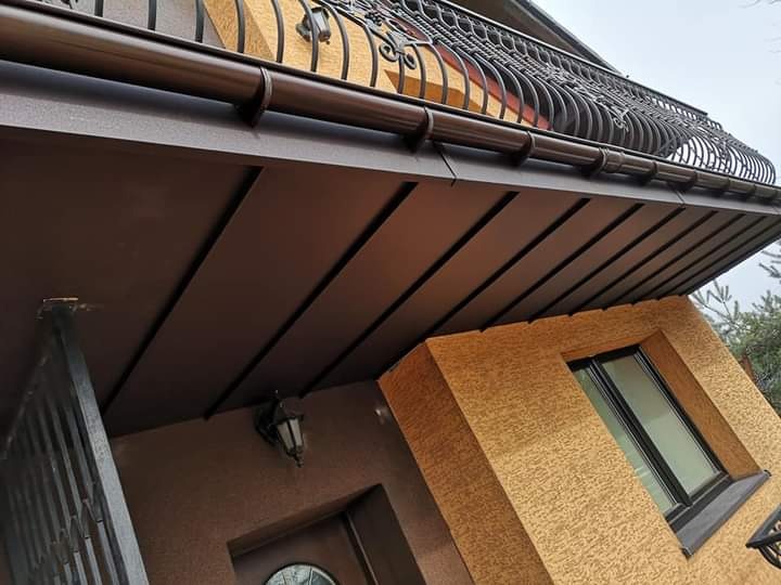 Ciemnobrązowe, metalowe orynnowanie domu z ozdobną, kutą balustradą na balkonie, widok z dołu na fragment elewacji z oknem.