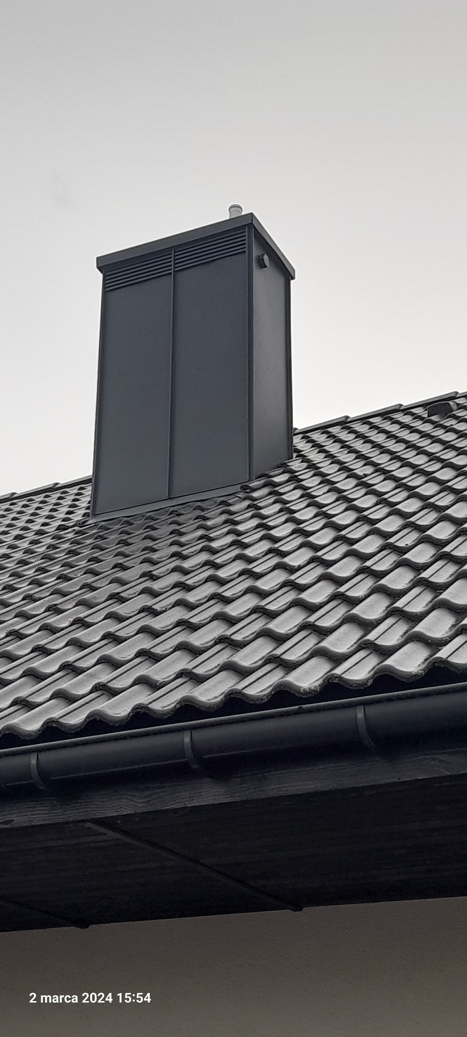 Szary komin wentylacyjny z blachy na dachu pokrytym dachówką ceramiczną w odcieniach szarości, widok z dołu na tle pochmurnego nieba.