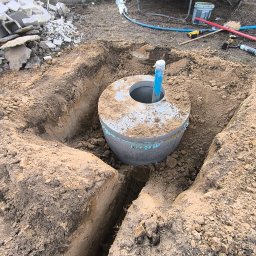 HYDRATOM TOMASZ ŻYHALUK - Betonowy krąg studzienny z niebieską rurą wystającą z otworu, umieszczony w wykopie ziemnym, widoczne narzędzia i gruz w tle.