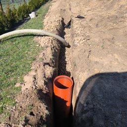 HYDRATOM TOMASZ ŻYHALUK - Wykop z pomarańczową rurą karbowaną w ziemi, wypełniony żwirem, biały wąż na powierzchni, w tle budynek i mężczyzna z rurą.