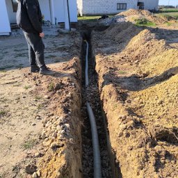 HYDRATOM TOMASZ ŻYHALUK - Wykop z dwiema rurami kanalizacyjnymi w ziemi, podsypany kamieniami, obok stoi osoba w kurtce z kapturem, widoczny fragment budynku mieszkalnego w tle.