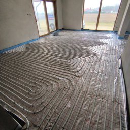 HYDRATOM TOMASZ ŻYHALUK - Białe rury ogrzewania podłogowego ułożone w pętle na folii z czerwoną siatką w jasnym, surowym wnętrzu z dużymi oknami i widokiem na zewnątrz.