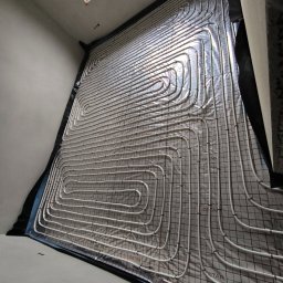 HYDRATOM TOMASZ ŻYHALUK - Białe rury ogrzewania podłogowego ułożone na izolacji z folią aluminiową i siatką, widok z góry pod kątem, w tle okno dachowe.