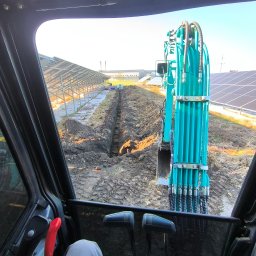 HYDRATOM TOMASZ ŻYHALUK - Widok z kabiny turkusowej koparki podczas wykonywania wykopu pod instalację paneli słonecznych, widoczne panele w tle.