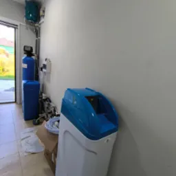 Biało-niebieski zmiękczacz wody stoi w rogu pomieszczenia, widoczne również elementy instalacji wodnej, rury i zawory, karton na podłodze.