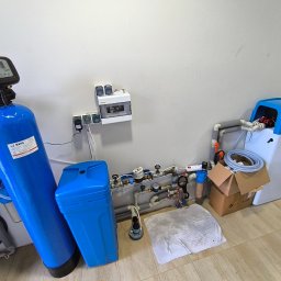HYDRATOM TOMASZ ŻYHALUK - Zestaw urządzeń do uzdatniania wody: duży, niebieski filtr kolumnowy z elektronicznym sterowaniem, obok mniejszy zbiornik, instalacja rurowa z zaworami, oraz zmiękczacz wody w obudowie...