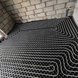 Białe rury grzewcze ułożone w regularnych pętlach na czarnej macie izolacyjnej, widoczne surowe ściany z cegieł i instalacja wodna w tle.