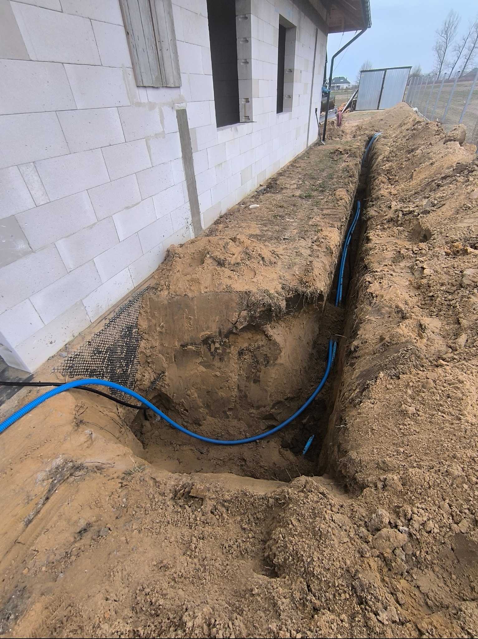 Wykop z ułożonymi niebieskimi rurami karbowanymi wzdłuż fundamentu budynku z białego bloczka, widoczna siatka drenażowa przy ścianie.