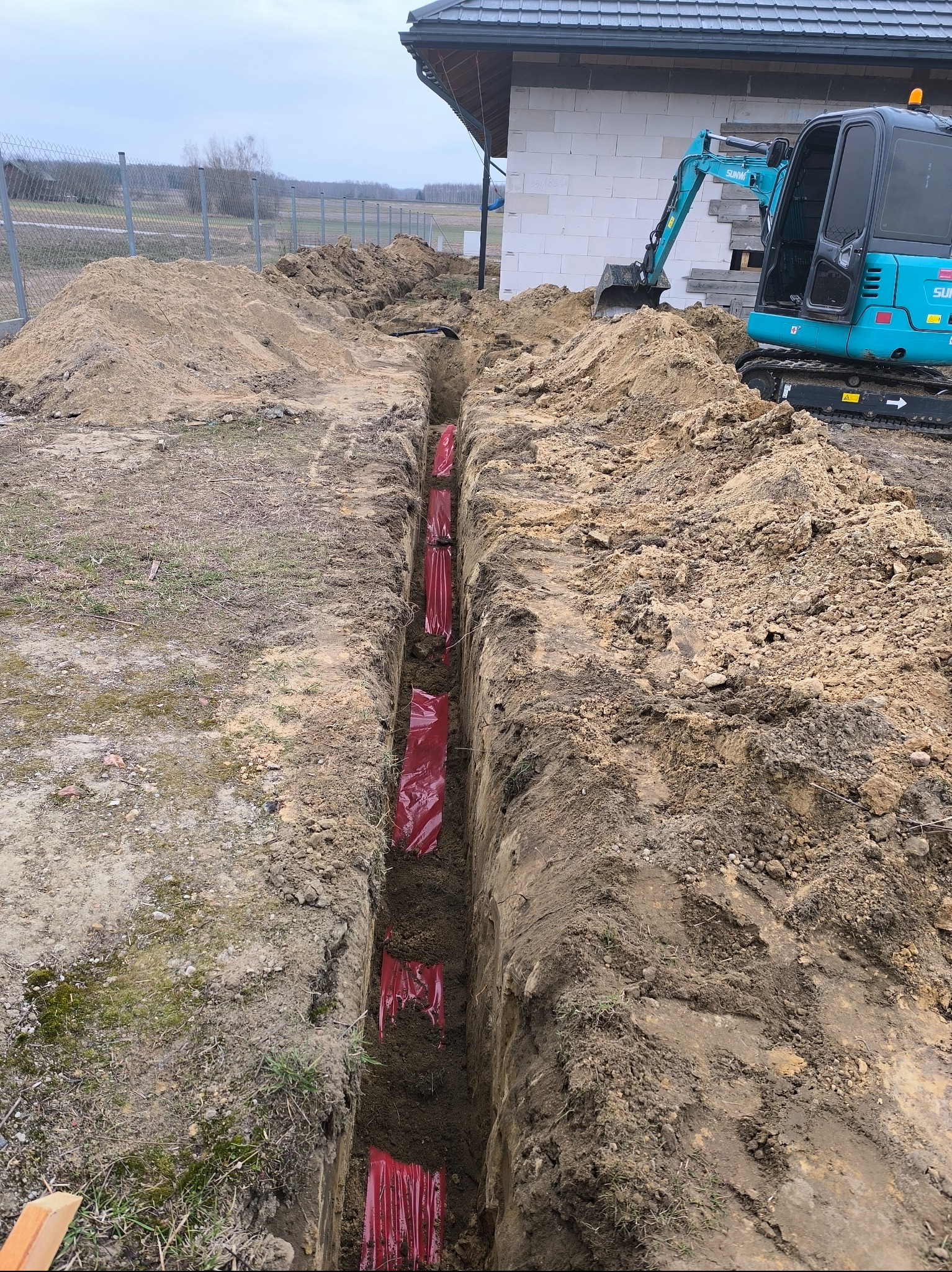 Wykop w ziemi z widocznymi czerwonymi rurami w folii, częściowo zasypany, z boku stoi mała koparka, w tle budynek w trakcie budowy z białego pustaka.