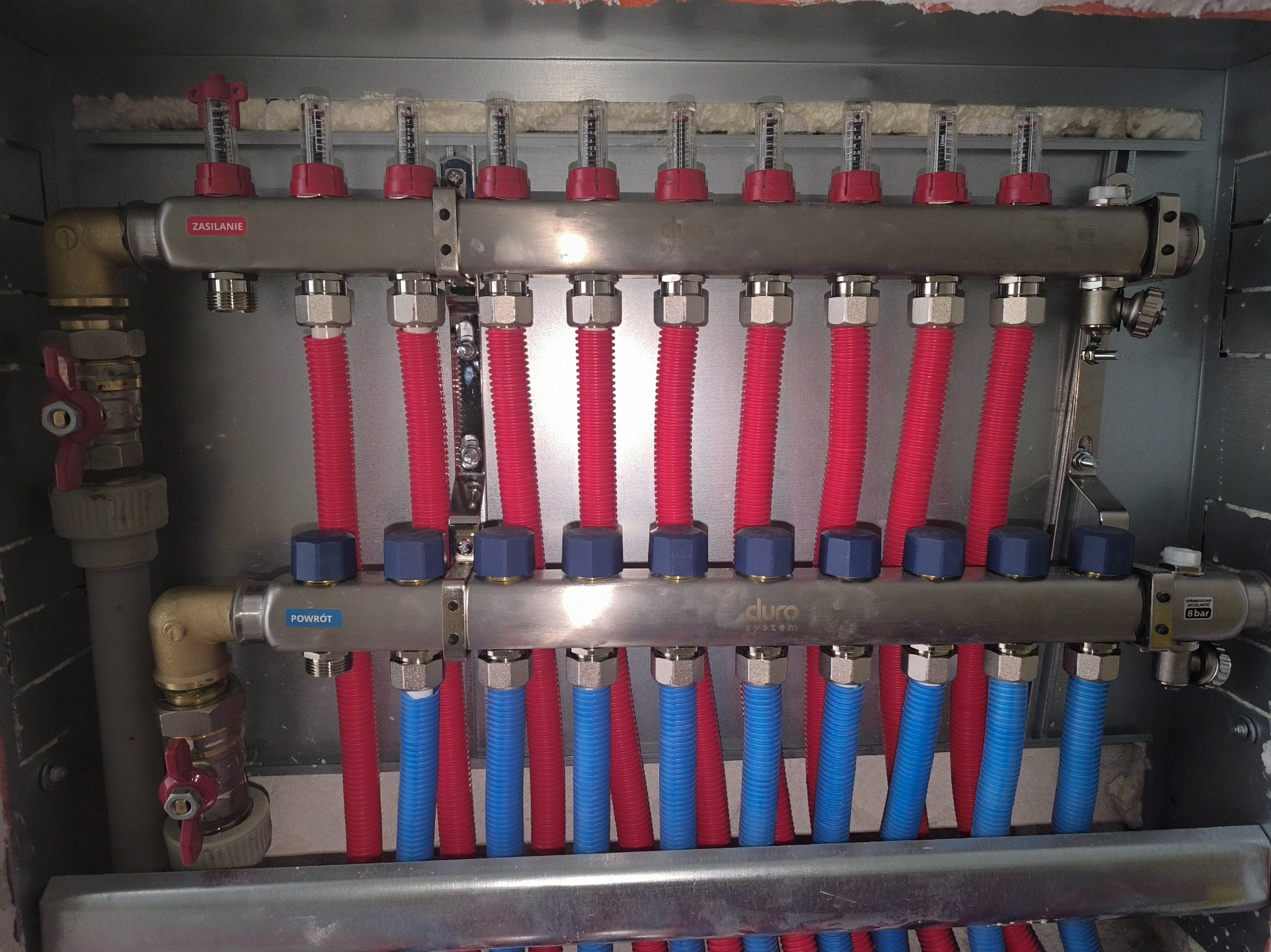 Rozdzielacz ogrzewania podłogowego z rurami w kolorach czerwonym i niebieskim, zamontowany w szarej szafce, widoczne etykiety 'Zasilanie' i 'Powrót'.