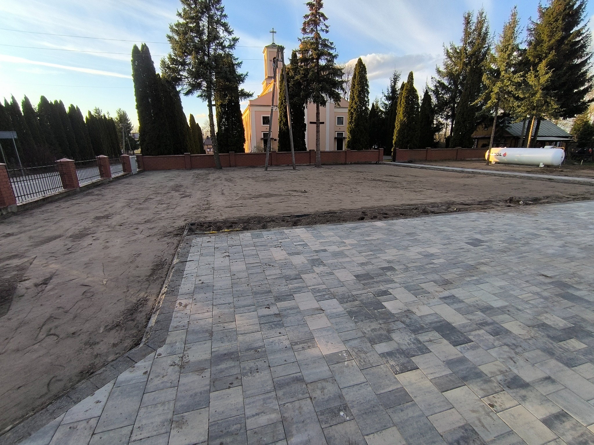 Ułożona szara kostka brukowa na placu budowy przed kościołem, widoczny zbiornik gazu płynnego w tle.
