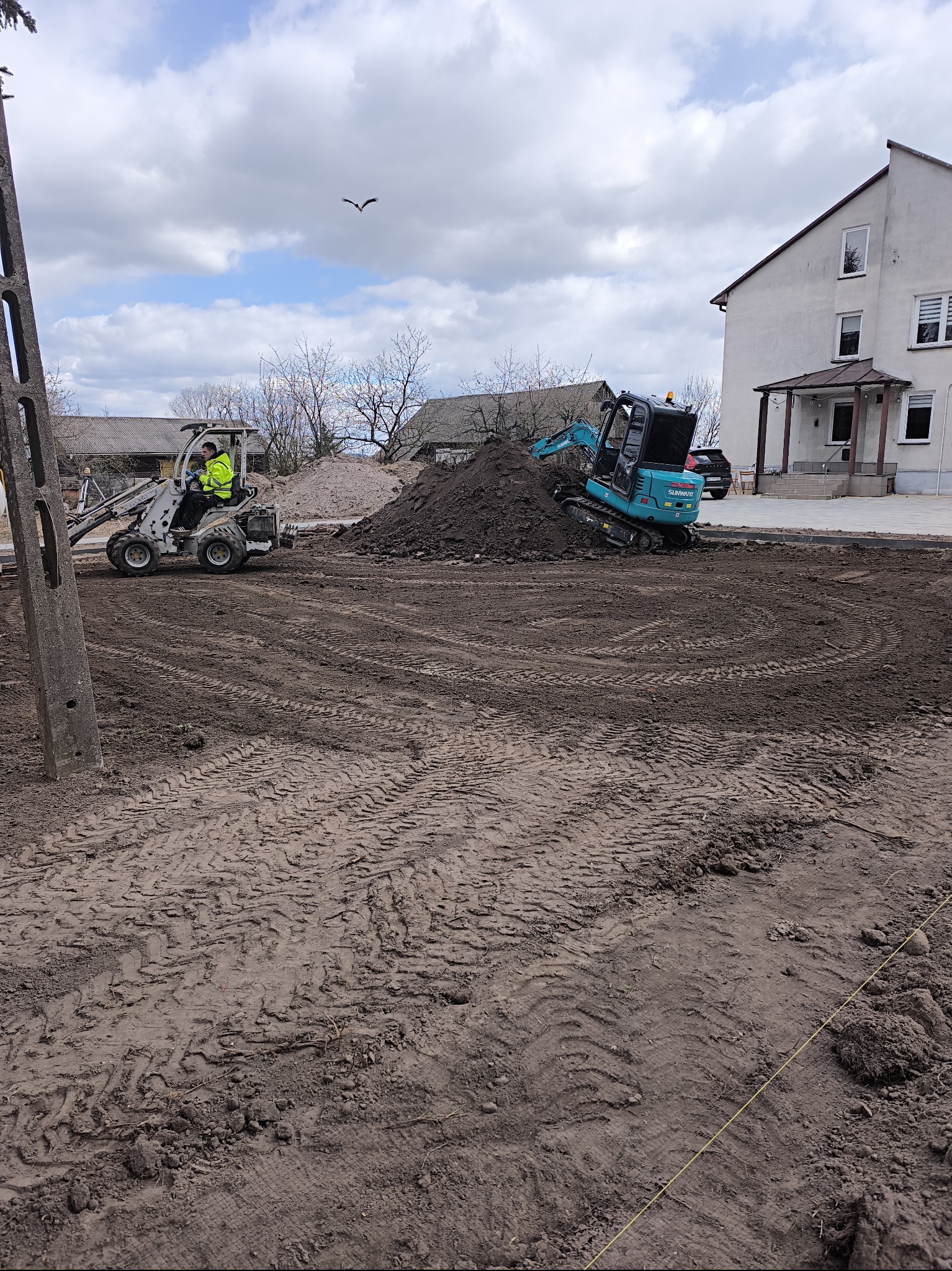 Przygotowanie terenu pod budowę przydomowej oczyszczalni ścieków: miniładowarka i minikoparka na placu budowy, widoczne ślady opon na ziemi i pryzma ziemi, w tle budynek mieszkalny.