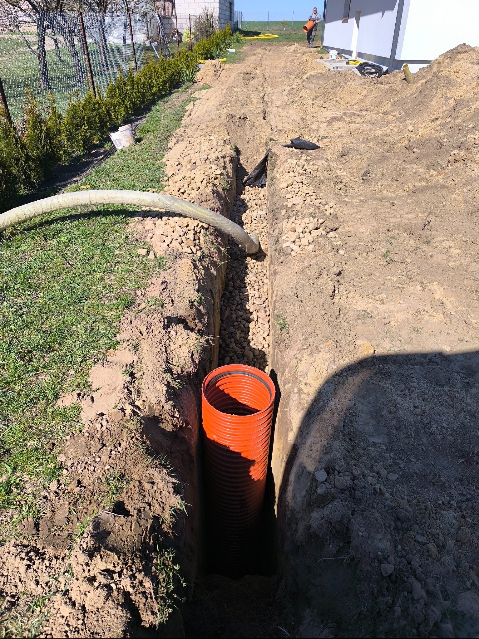 Wykop z pomarańczową rurą karbowaną w ziemi, wypełniony żwirem, biały wąż na powierzchni, w tle budynek i mężczyzna z rurą.