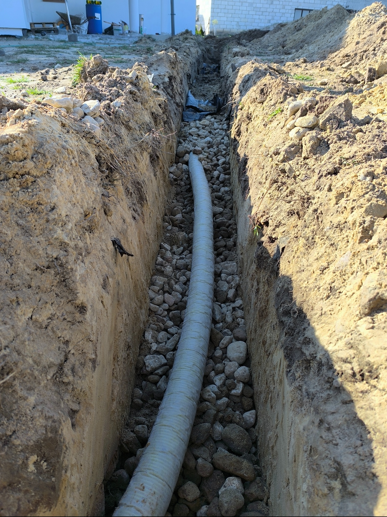 Wykop w ziemi z ułożoną rurą drenarską i kamieniami, widoczne ślady korzeni w ścianach wykopu, w tle budynek w trakcie budowy.