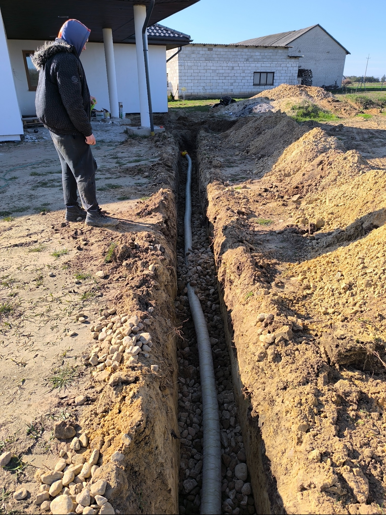 Wykop z dwiema rurami kanalizacyjnymi w ziemi, podsypany kamieniami, obok stoi osoba w kurtce z kapturem, widoczny fragment budynku mieszkalnego w tle.