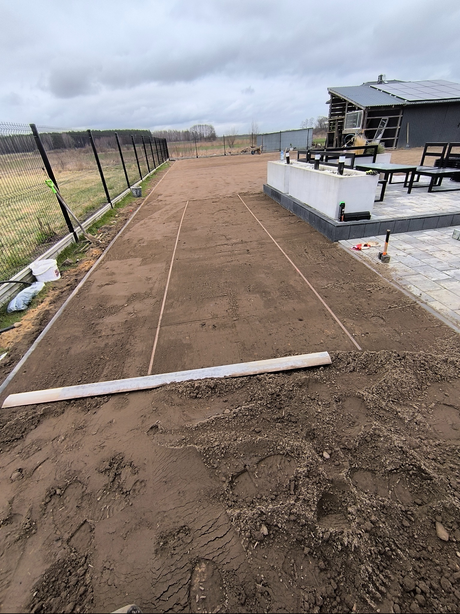 Przygotowanie terenu pod budowę ścieżki lub tarasu: wyrównana ziemia z wyznaczonymi liniami, widoczne narzędzia i materiały budowlane, w tle ogrodzenie i budynek z panelami słonecznymi.