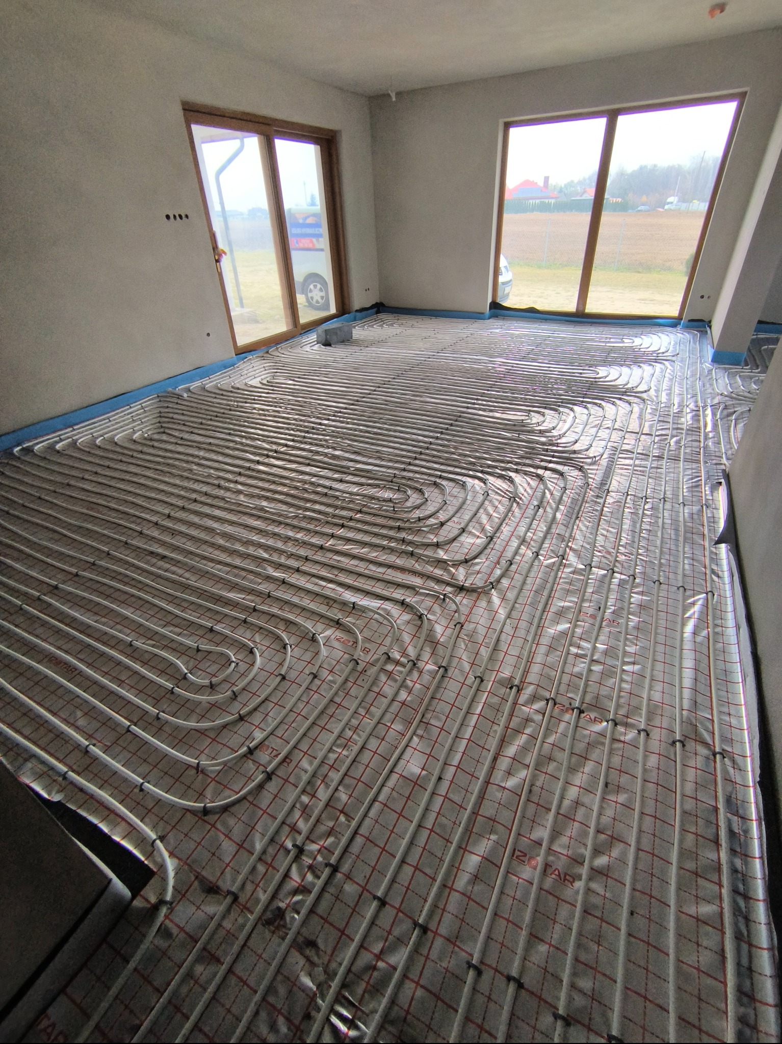 Białe rury ogrzewania podłogowego ułożone w pętle na folii z czerwoną siatką w jasnym, surowym wnętrzu z dużymi oknami i widokiem na zewnątrz.