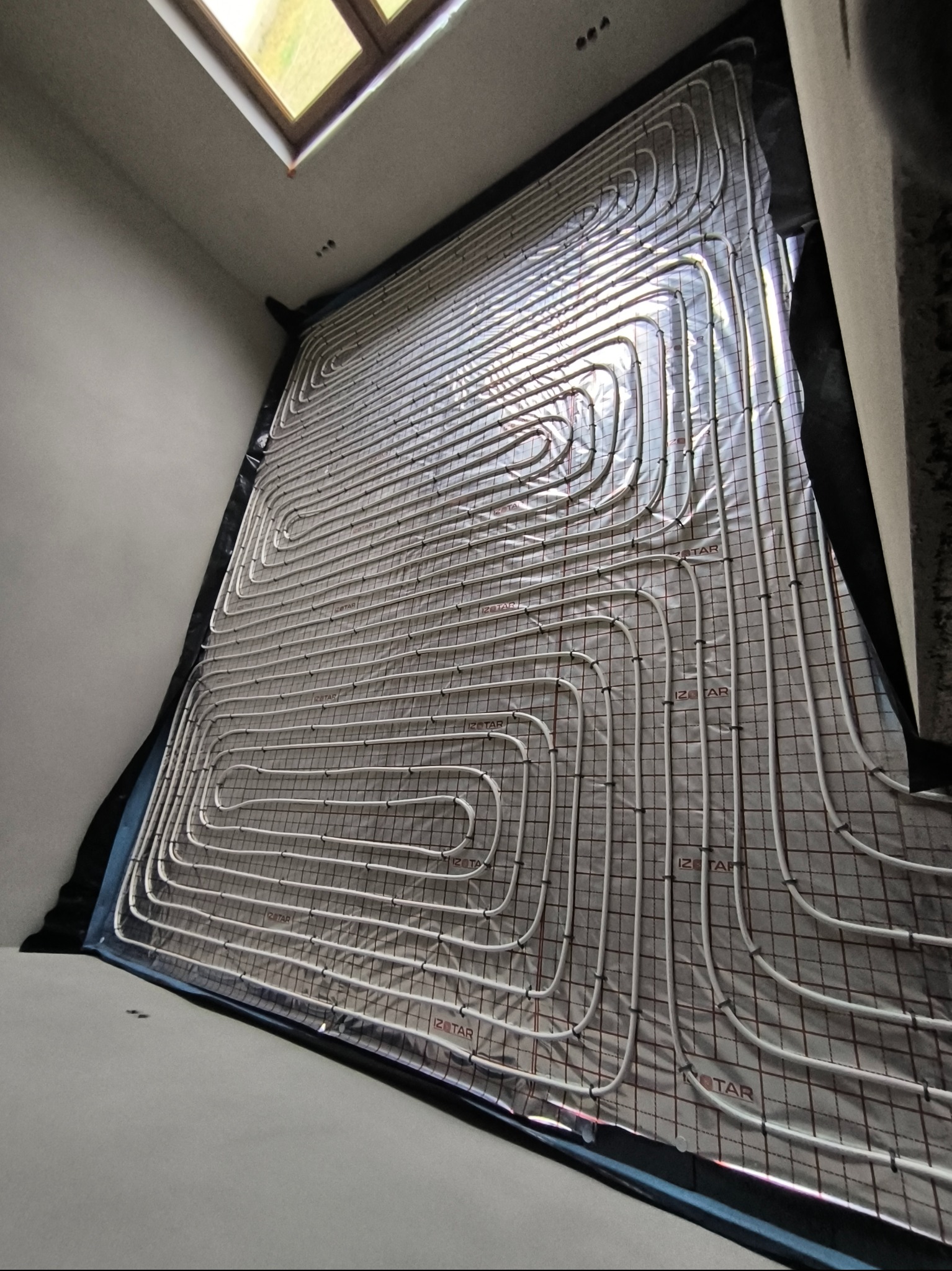 Białe rury ogrzewania podłogowego ułożone na izolacji z folią aluminiową i siatką, widok z góry pod kątem, w tle okno dachowe.