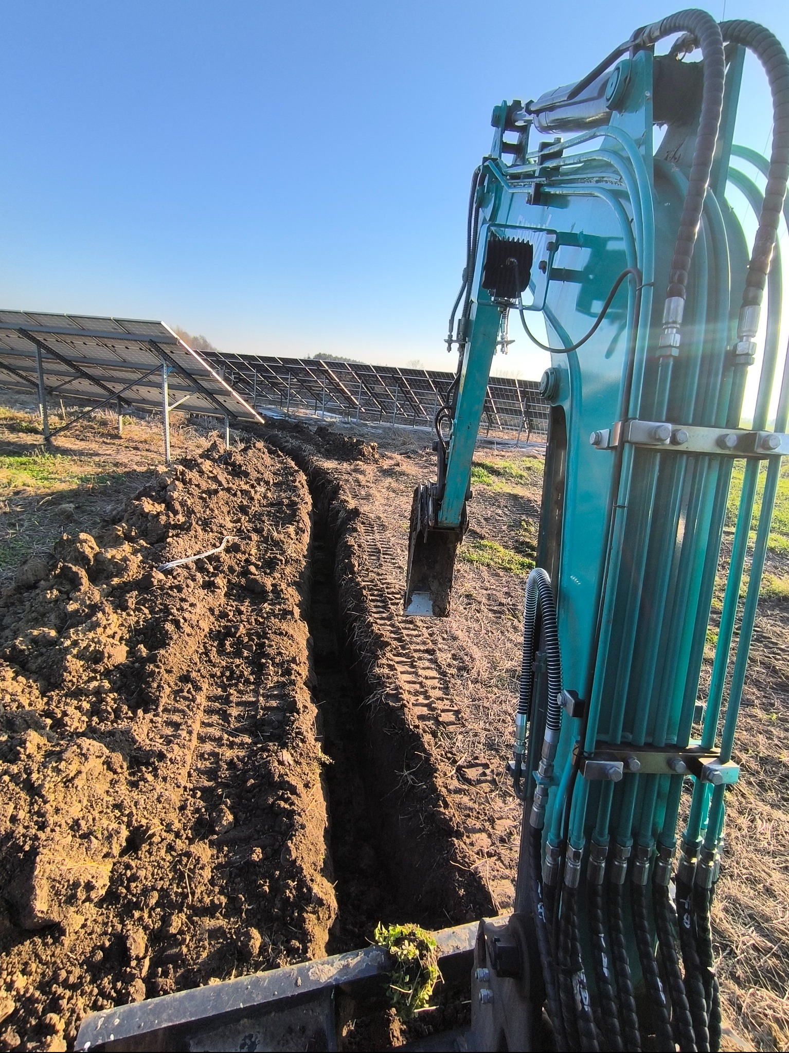 Turkusowa minikoparka wykopuje wąski rów w ziemi, w tle panele słoneczne pod jasnym niebem.