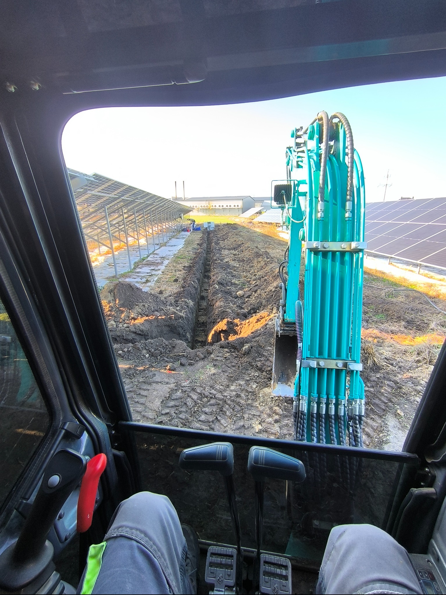 Widok z kabiny turkusowej koparki podczas wykonywania wykopu pod instalację paneli słonecznych, widoczne panele w tle.